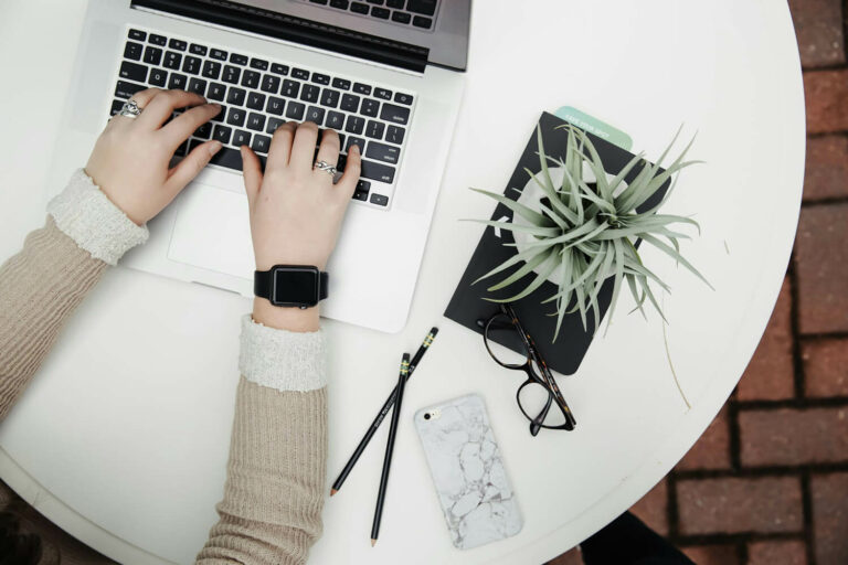 personne tapant sur le clavier d'un ordinateur portable, sur une table ronde blanche. A côté sont posés des lunettes, une plante et un téléphone.