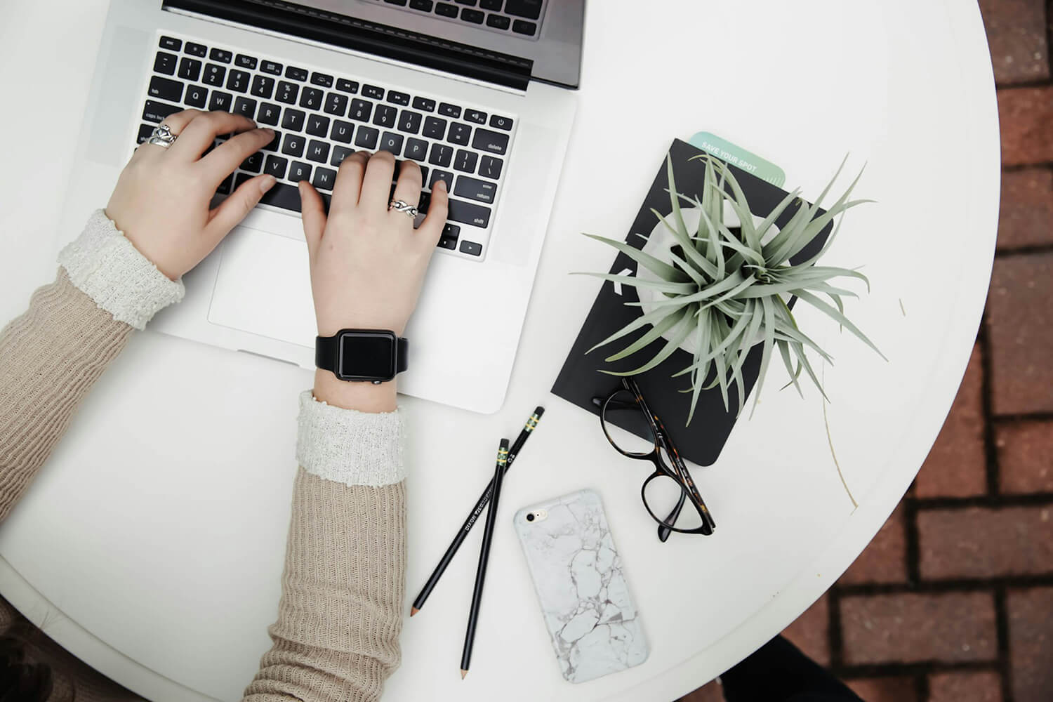 personne tapant sur le clavier d'un ordinateur portable, sur une table ronde blanche. A côté sont posés des lunettes, une plante et un téléphone.